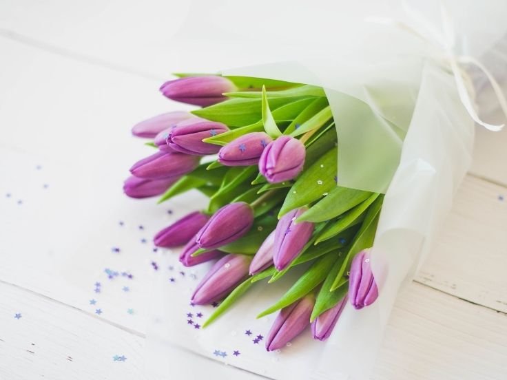 A bouquet of fresh purple tulips wrapped in white paper, placed on a white wooden surface with scattered small star-shaped confetti.